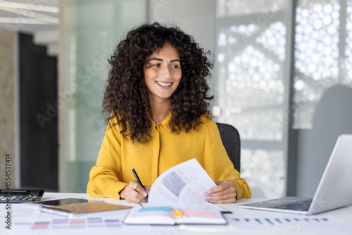 A smiling businesswoman in a yellow shirt reviews documents at her desk in a modern office setting, working with a laptop and other office supplies.