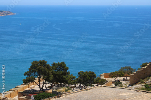 Mediterranean Sea View from Santa Bárbara Castle in Alicante with Stone Walls, Pine Trees, and Azure Water under Clear Blue Sky