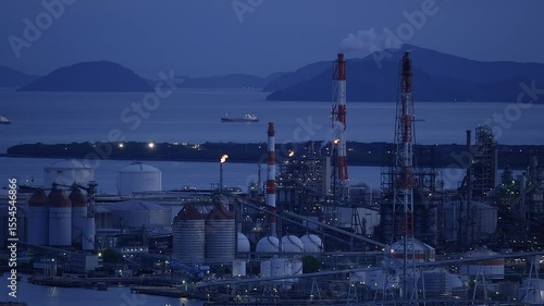 A sprawling industrial plant with smoking chimneys and burning flare stacks at dusk.