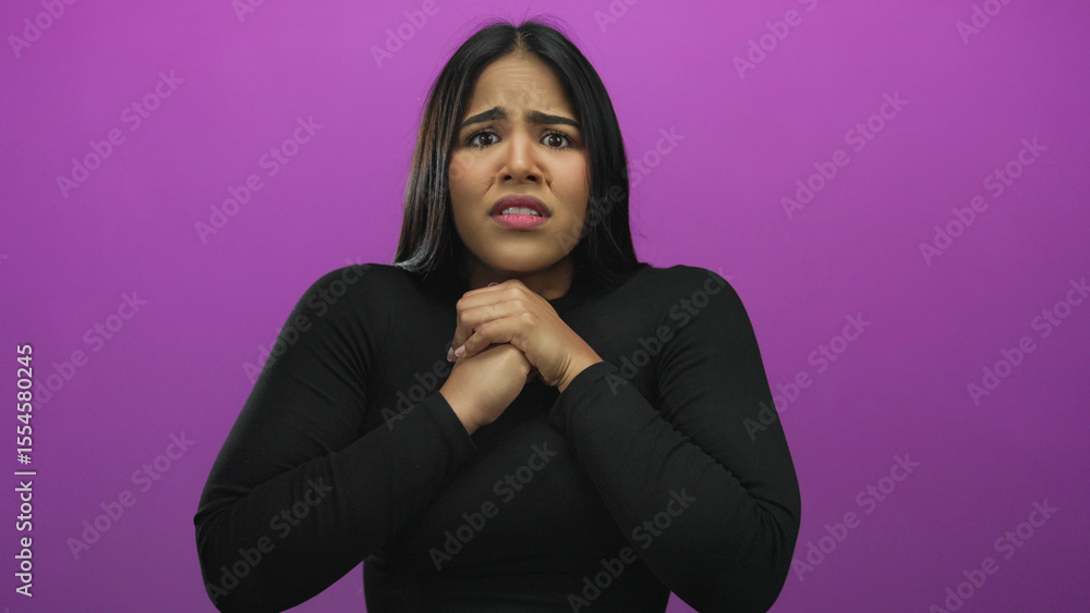 Fototapeta premium Young woman with dark hair and concerned expression, clasping hands on pink background, conveying anxiety and stress.