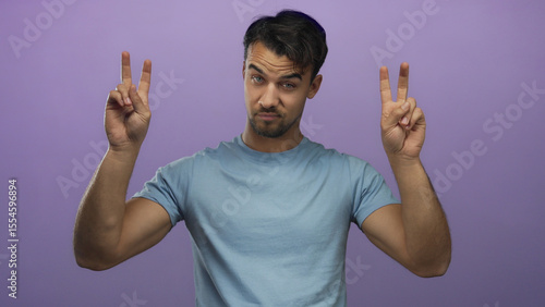 Young hispanic man wearing blue shirt makes air quotes gesture against an isolated purple background, expressing sarcasm or irony in a humorous and engaging manner.