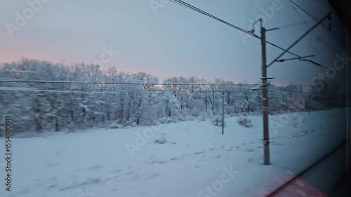 Train window shows a snowy landscape with trees and wires. The sky is dark blue and the sun is setting. view from moving train commuter transport passenger express.