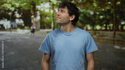 Handsome hispanic man in blue shirt gazes upwards in a peaceful green park surrounded by trees and natural greenery on a sunny day
