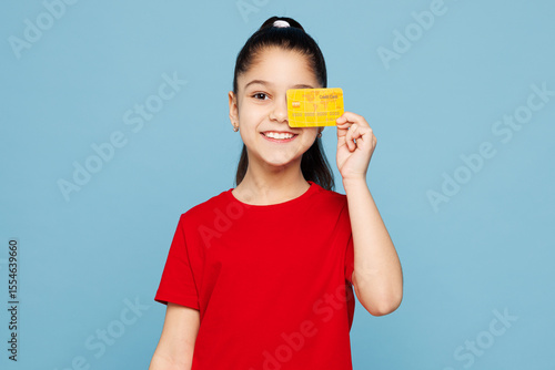 Fotografie Little kid girl 8-9 years old wears red t-shirt hold in hand cover eye with mock up of credit bank card isolated on blue background child studio portrait