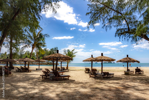 Klong Prao Beach with rustic thatched parasols shading wooden sunbeds under palms, leading to turquoise waves and island-dotted horizon. Koh Chang, Thailand.


