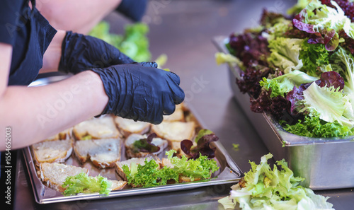 Hands in black gloves preparing healthy sandwiches with fresh lettuce in a professional kitchen. Food hygiene, catering service, or restaurant prep concept.