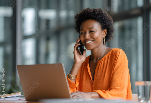Woman with short hair smiles while using a laptop and talking on a cell phone