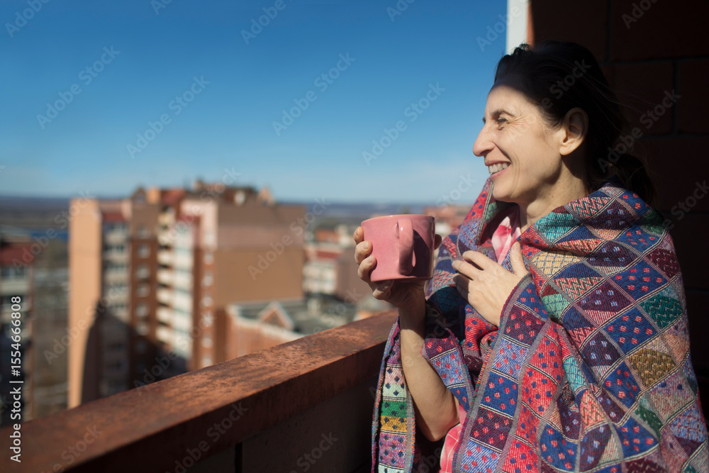 Fototapeta premium happy middle aged woman taking cover with a blanket drinking tea on the balcony of your apartment on a sunny spring day. Slow life. Enjoying the little things.