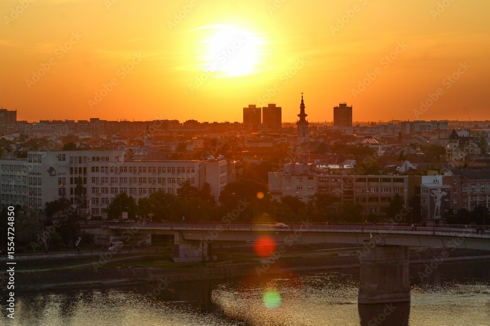 Fototapeta premium Beautiful sunset over Novi Sad, Serbia, with city buildings, a church tower, and a bridge reflected in the calm river below.
