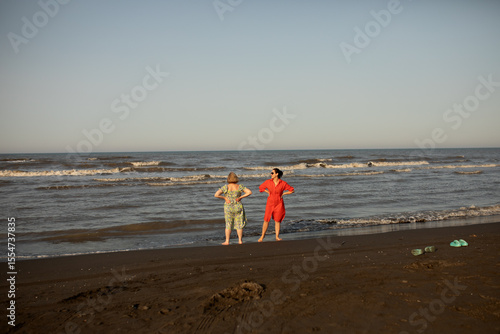 Two women enjoying a joyful seaside moment during vacation, barefoot on the beach, sharing laughter and sunshine. A memory of freedom, travel, and connection