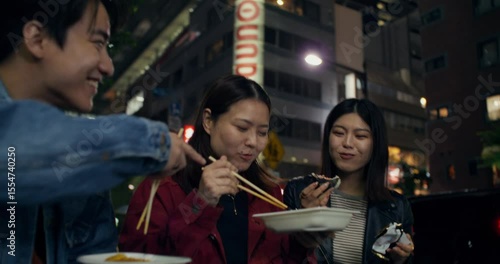 Three young people are eating street food and talking while standing in the night city. They laugh and look happy. Tokyo, Japan
