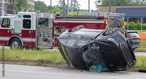 A crashed automobile had turned over and is laying on its side with the windshield smashed with a fire truck on the scene
