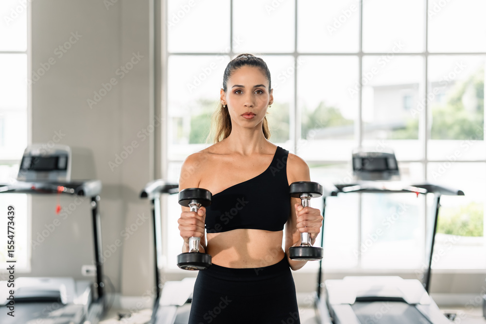 Fototapeta premium A woman is holding two dumbbells in her hands. She is standing in a gym. The dumbbells are black and silver