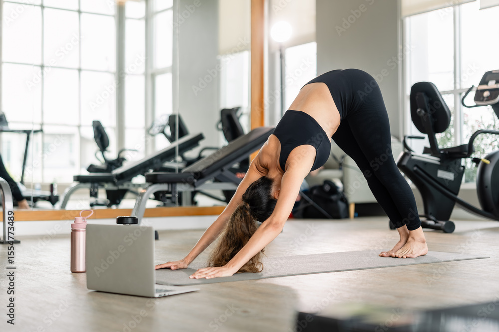 Fototapeta premium A woman is doing a yoga pose on a mat. A laptop is on the mat next to her