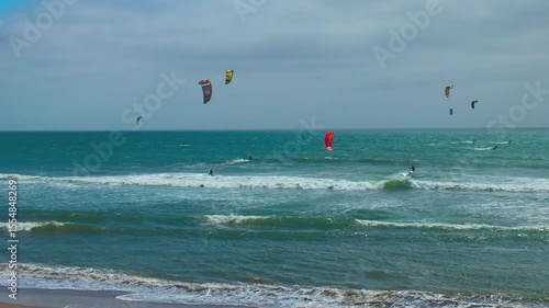 Kitesurfers ride ocean waves near Pacifica, California, along Highway 1. Colorful kites fill the cloudy sky over the turquoise sea in this action-packed coastal scene