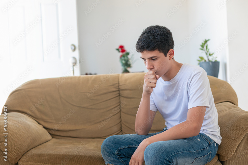 A young man sits on a couch, covering his mouth with his hand A young man sits on a couch, covering his mouth with his hand