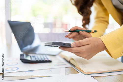 A businesswoman is working in her office. She is checking documents at her desk with a calculator and a mobile phone. She is looking at the calculator and thinking.

