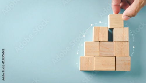A hand stacks wooden blocks to form a pyramid, symbolizing growth, strategy, and building foundations against a light blue background.