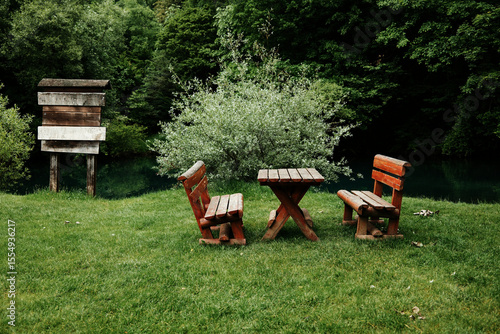 Obraz na plátně Wooden benches and a table stand on green grass by a calm river, surrounded by dense trees