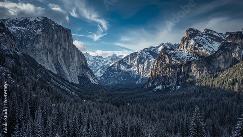 A scenic view of a valley with snow capped mountains and a dense forest under a cloudy blue sky