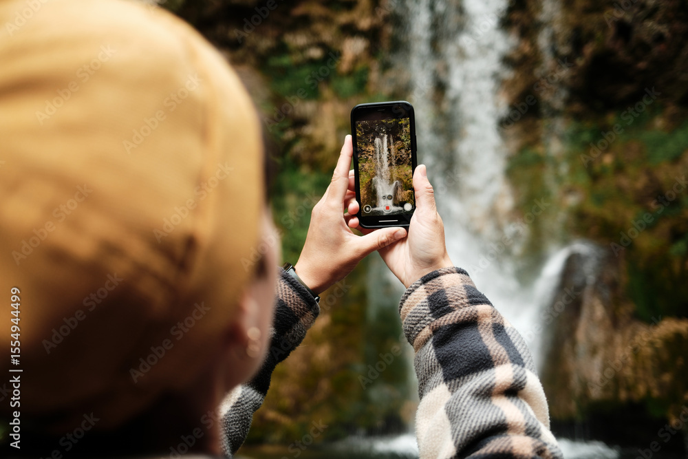 Fototapeta premium Female tourist in plaid jacket filming Veliki Buk waterfall with smartphone at Lisine reserve, Serbia. Travel moment captured in nature. Travel blogger making video outdoor. Close back view.