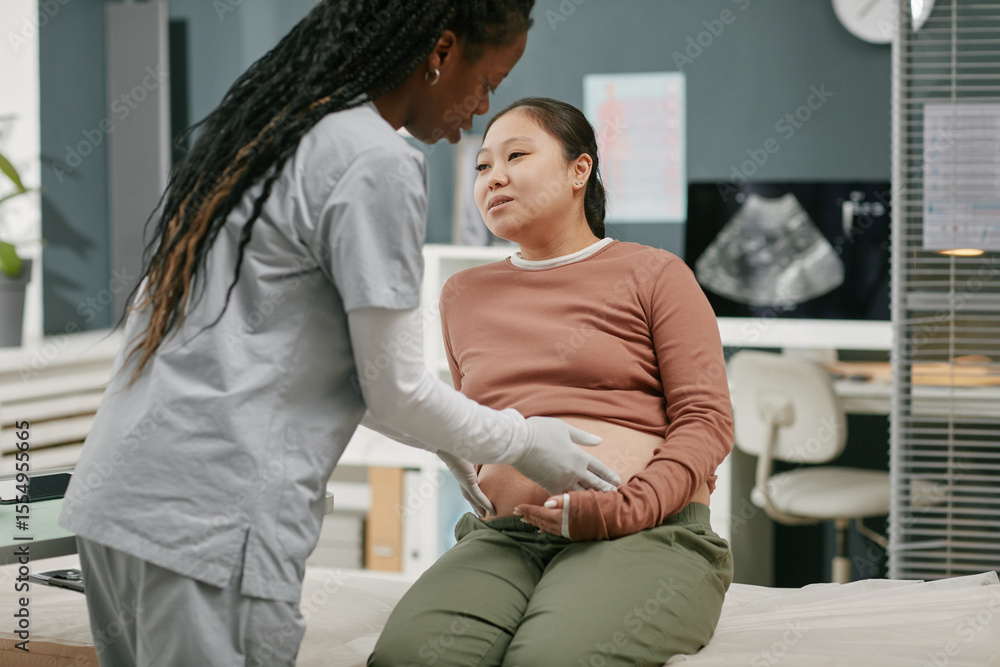 Fototapeta premium Asian pregnant young adult woman sitting on examination table interacting with Black woman healthcare professional during prenatal checkup in medical clinic setting