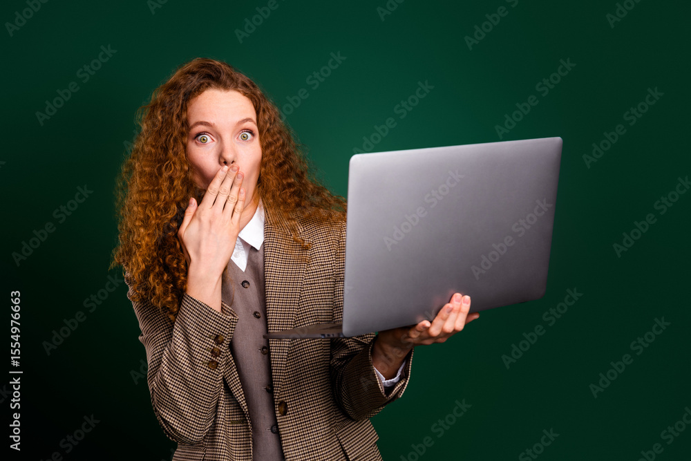 Naklejka premium Young surprised businesswoman with red curly hair using a laptop, wearing elegant suit, standing against green background