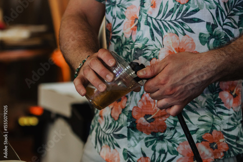 Close-up of two hands pouring draft beer from a tap into a plastic cup, featuring one hand in a floral Hawaiian shirt and green beaded bracelet against a blurred bar backdrop