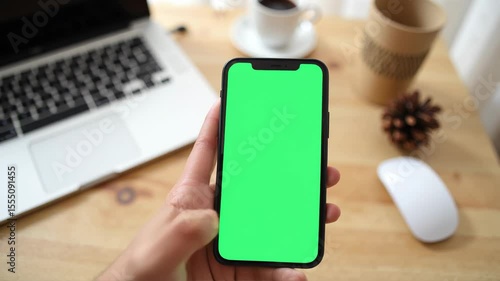 Hand holding smartphone green screen mockup on office desk with laptop coffee technology business work online concept