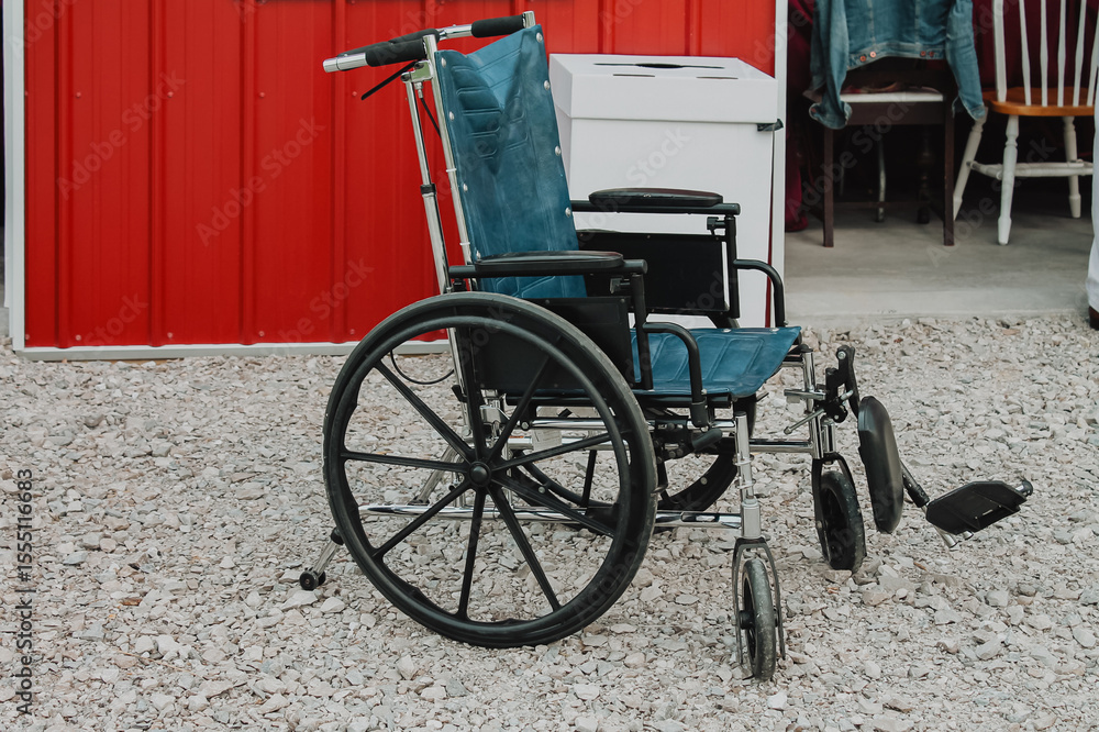 Fototapeta premium Manual wheelchair parked on gravel outside a red metal-sided barn entrance with chairs draped in a denim jacket visible inside