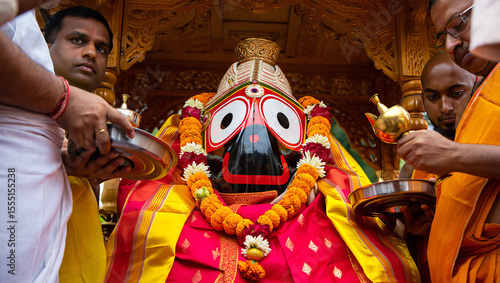 A close-up photograph captures the Jagannath deity statue inside an ornate wooden chariot, with the deity's painted wooden face showing distinctive large round eyes and traditional features