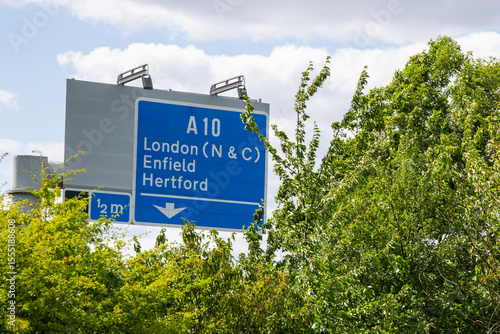 M25 motorway road sign for A10 slip road to London (N & C), Enfield, and Hertford, photographed through roadside foliage in summer.