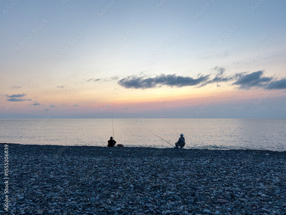Fototapeta premium Two people fishing on pebble beach at sunset, peaceful sea view