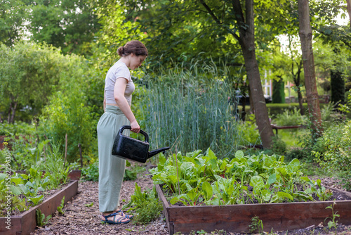 Young woman watering vegetables in raised garden bed using watering can. Sustainable gardening, healthy living, eco-friendly backyard, slow life and organic food concept. Copyspace