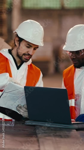 Two construction workers in safety gear analyze plans together, focusing intently on their computer in a bustling worksite full of activity