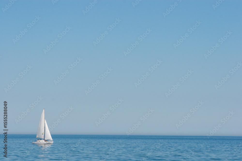 Obraz premium A single white sailboat gracefully glides across the calm, blue expanse of the sea under a clear, vast sky. Perfect for themes of freedom, tranquility, travel, and adventure on water