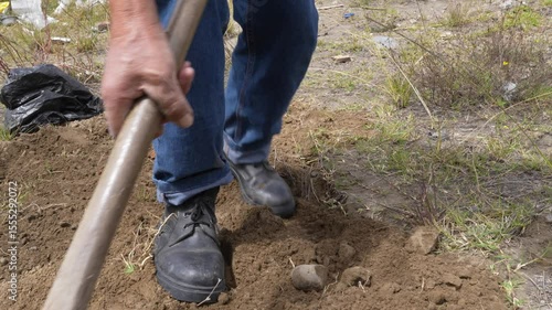 Gravedigger wearing blue jeans and black shoes covering burial site with pickaxe, manually filling ground with loose earth in rural Ecuadorian cemetery during daytime
