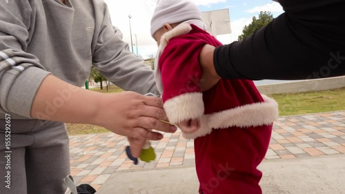 Adorable baby boy wearing Santa Claus costume practicing his first steps with the help of mom and aunt in a park