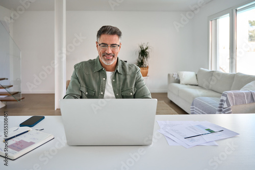 Tableau sur toile Smiling happy older mature middle aged professional man wearing eyeglasses looking at computer sitting at living room table, using laptop hybrid working, paying bills, browsing online at casual home
