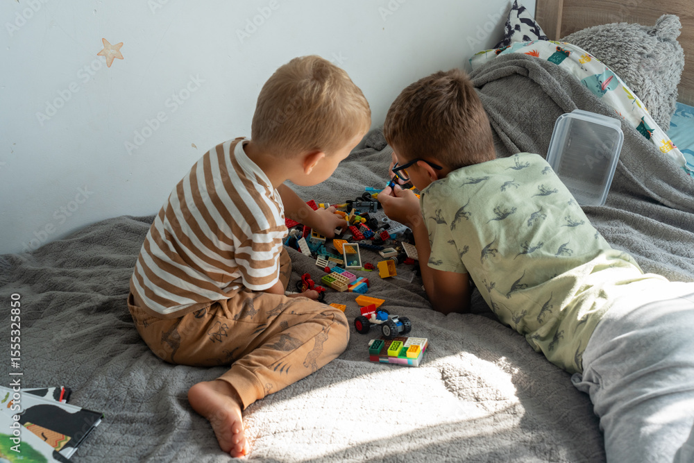 Naklejka premium Two little boys playing with colorful plastic building blocks on cozy bed in sunlit kids bedroom. Concept of childhood creativity, family bonding, indoor play and educational toys.