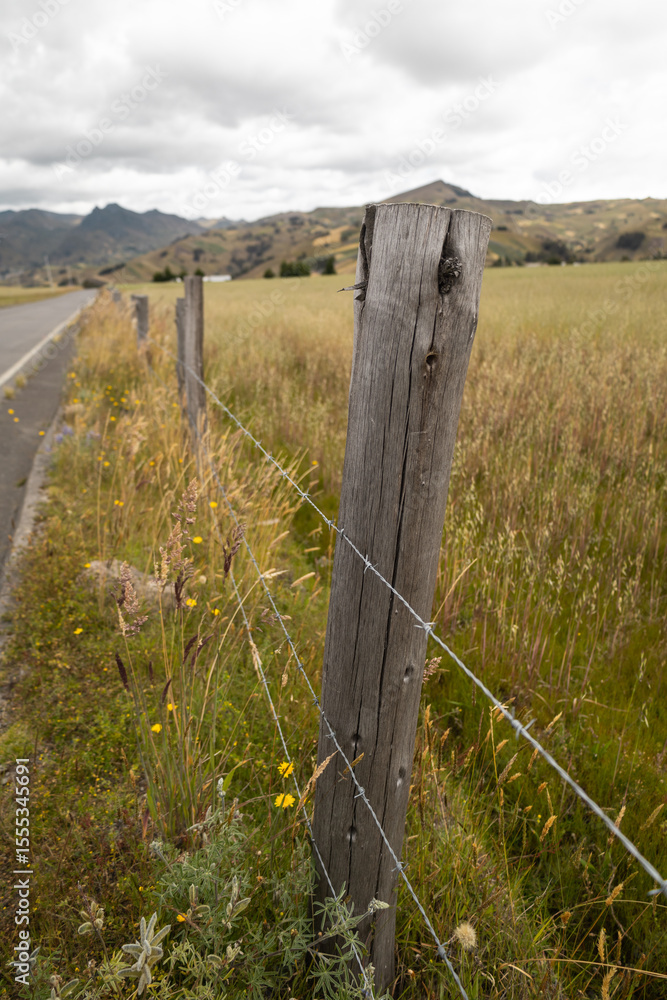Fototapeta premium From a moving car, a wooden fence post with barbed wire marks the boundary of a rural field. The view captures rustic charm, open space, and the quiet of country life.