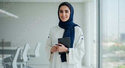 Confident Emirati businesswoman smiles, holding a tablet and pen in a modern office.