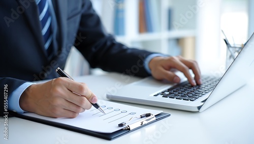 A detail-oriented businessman in a formal suit meticulously manages his workflow by writing on a checklist while concurrently operating a laptop, showcasing professional multitasking at his desk.