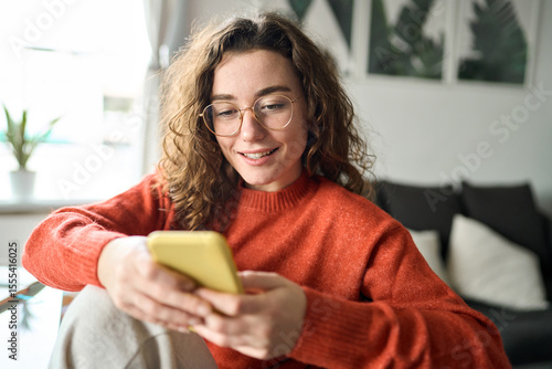Happy girl checking cell phone online education apps, texting, browsing internet for shopping sitting at home. Young smiling woman wearing glasses holding smartphone using cellphone, looking at mobile