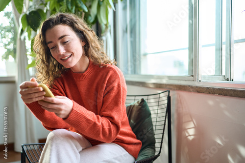 Slika na platnu Happy young woman sitting on chair holding mobile phone using cellphone device, looking at smartphone, checking modern apps, texting messages, browsing internet doing shopping relaxing at home