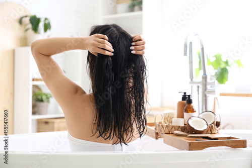 Beautiful young woman applying hair mask with coconuts in bathroom at home, b...