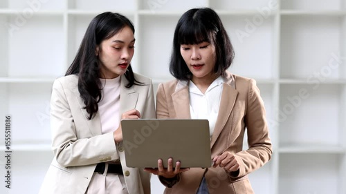 Professional businesswoman working collaboratively on laptop, strategizing and problem-solving together, corporate office while standing near window.