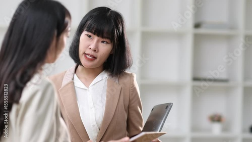 Professional Asian businesswoman collaborating in office, holding clipboard and folder while discussing work details and shared strategic approach.