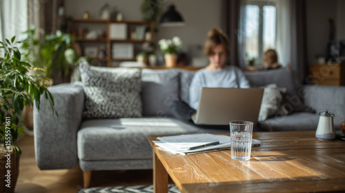 White woman working on laptop on sofa with child in background, wooden coffee table with glass of water and papers, bright home interior with plants, casual outfit