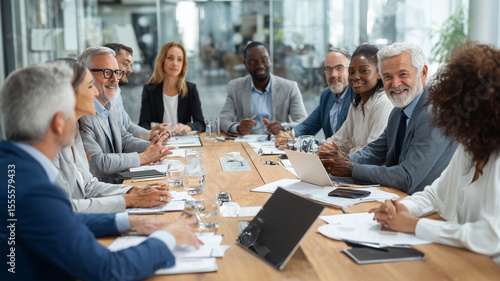 Large group of racially diverse senior and middle-aged business professionals in formal attire seated around a wooden conference table in a bright glass-walled office during daytime